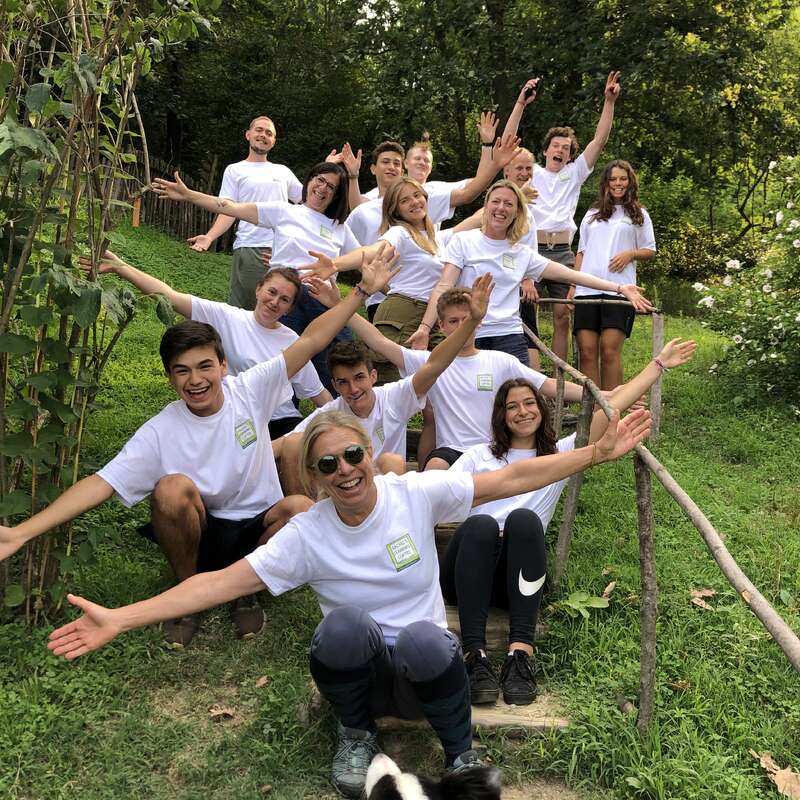 La imagen muestra a un grupo de 14 personas posando en una escalera de madera en una frondosa zona de césped, con un perro en primer plano. Llevan camisetas blancas a juego.