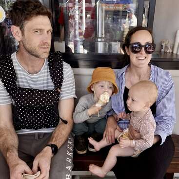 Uma família de quatro pessoas está sentada em um banco. O pai parece sério, enquanto a mãe, usando óculos escuros, sorri. Seus dois filhos pequenos gostam de tomar sorvete.