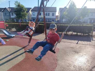 Um menino e uma menina se divertem balançando em um playground em um dia ensolarado, cercados por casas, árvores e um cenário suburbano iluminado.