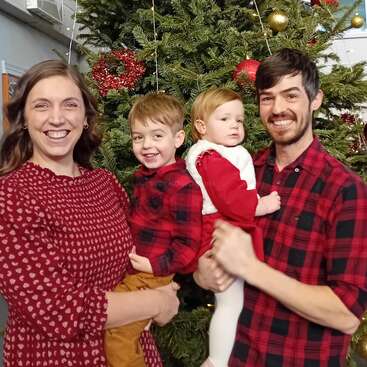 A imagem mostra uma família de quatro pessoas posando em frente a uma árvore de Natal decorada, com a mãe e o pai segurando seus dois filhos. A família está vestida com roupas vermelhas e brancas.