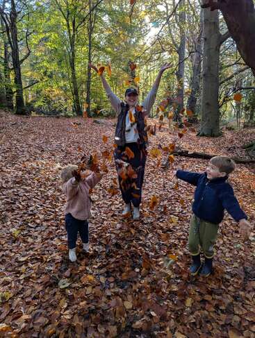 Uma mulher e duas crianças brincam alegremente nas folhas de outono em uma floresta iluminada pelo sol, jogando folhas no ar e sorrindo, cercadas pelas cores vibrantes do outono.