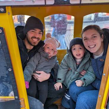 A family of four, bundled in coats and hats, sits happily inside a yellow vehicle. Parents smile while holding their two young children, enjoying a cozy moment.