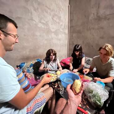 A imagem mostra um grupo de pessoas reunidas em torno de uma mesa, descascando legumes e preparando alimentos em um ambiente aconchegante e rústico.
