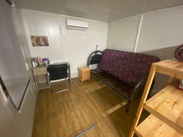 A small, tidy room with wooden flooring, a purple futon, desk and chair, air conditioner, small nightstand, shelving unit, and a wall picture. Cozy atmosphere.