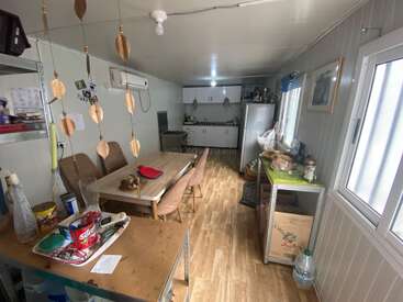 This image shows a cozy kitchen and dining area with a wooden table, chairs, shelves, refrigerator, hanging decorations, window, and various household items scattered throughout.
