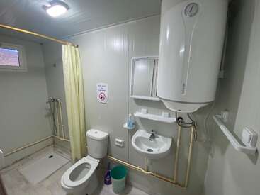 This image shows a simple bathroom with a toilet, sink, shower, mirror, water heater, yellow shower curtain, green trash bin, and a warning sign on the wall.