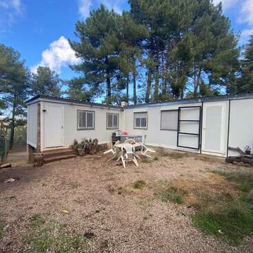 A small, white modular building stands in a wooded area. Plastic chairs and a table are outside on a dirt ground, surrounded by trees and nature.