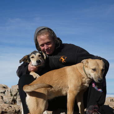 A person wearing a black hoodie crouches outdoors, embracing two brown dogs. The sky is clear and blue, with rocky terrain visible in the background.