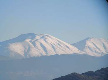 This image displays a beautiful snow-covered mountain range beneath a clear blue sky, with rugged dark hilltops visible in the foreground, creating a serene natural scene.