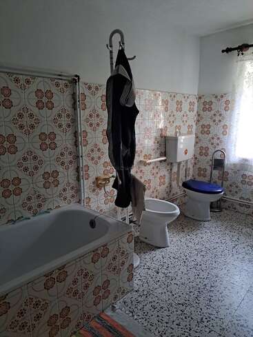 Esta casa de banho tem azulejos florais vintage, uma banheira, um vaso sanitário, um bidê, piso estampado, um cabide, uma janela com cortinas de renda e uma tampa de vaso sanitário azul.