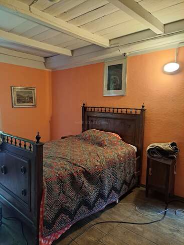 Um quarto aconchegante com uma cama antiga de madeira, colcha colorida, paredes laranja, teto de madeira, duas fotos emolduradas, uma mesinha de cabeceira, um abajur e piso de madeira.