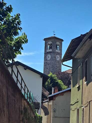 Uma torre do relógio de pedra ergue-se acima das casas pitorescas da vila, rodeada por vegetação. O céu azul brilhante e a luz do sol criam um cenário encantador, tranquilo e pitoresco de uma vila europeia.