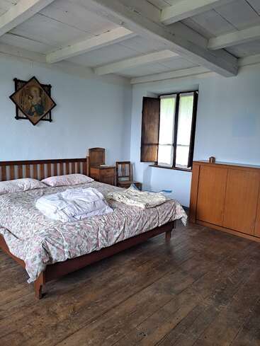 Este quarto aconchegante tem uma cama de madeira com roupa de cama com motivos florais, móveis rústicos de madeira, uma janela aberta, piso de madeira, vigas brancas no teto e uma pintura religiosa na parede.
