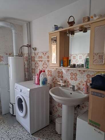 Esta imagem mostra um banheiro com azulejos com padrões retrô, uma máquina de lavar, um aquecedor de água, um lavatório com pedestal, um armário com espelho, produtos de limpeza e vários itens de higiene pessoal bem organizados.