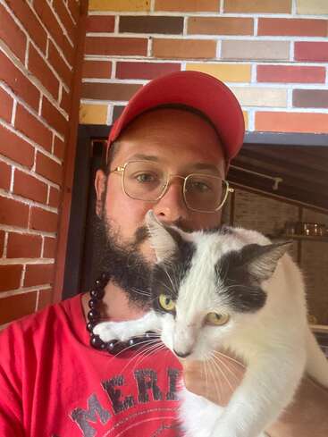 Ein Mann mit Brille, roter Mütze und rotem Hemd hält eine schwarz-weiße Katze im Arm. Sie stehen vor einer Backsteinmauer.