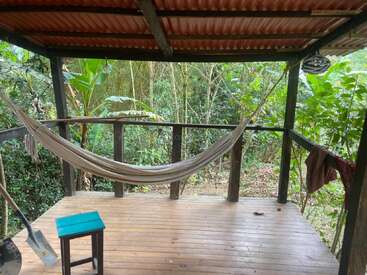 Eine ruhige Holzveranda mit einer Hängematte, einem kleinen blauen Hocker und einer Schaufel. Umgeben von üppigem, grünem Wald, der einen ruhigen, naturverbundenen Ort der Entspannung schafft.