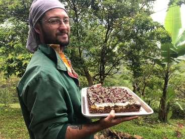Eine Person mit Brille und Bandana hält ein Tablett mit einer großen, verzierten Torte in der Hand. Sie ist umgeben von üppig grünen Bäumen und einer natürlichen Umgebung im Freien.