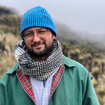 A man with glasses, a blue knit hat, and a black-and-white scarf smiles outdoors, wearing a green jacket. Foggy, grassy background with blurred nature scenery.