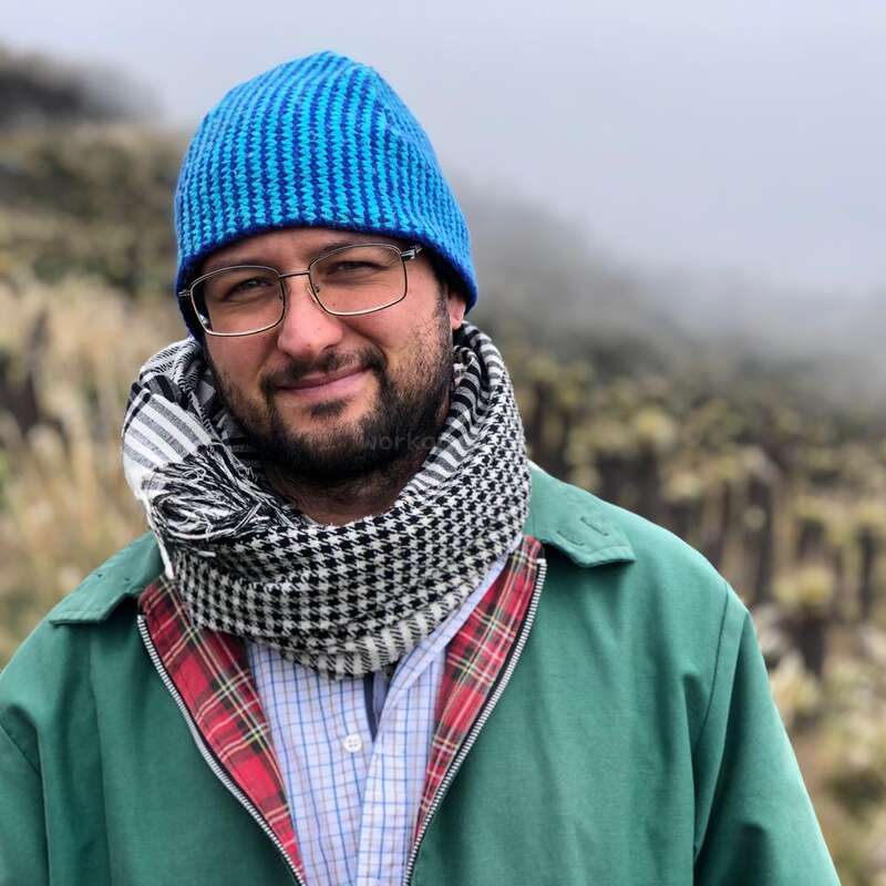 Ein Mann mit Brille, blauer Strickmütze und schwarz-weißem Schal lächelt draußen in einer grünen Jacke. Im Hintergrund sieht man nebliges Grasland mit verschwommenen Naturmotiven.