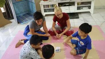 Cuatro niños y dos adultos están sentados en una suave alfombra, reunidos en círculo, jugando a las cartas juntos en una acogedora y luminosa sala de estar.