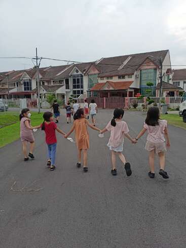 Un grupo de niños camina de la mano por una calle suburbana, sonriendo y jugando, con casas y vegetación a ambos lados bajo un cielo nublado.