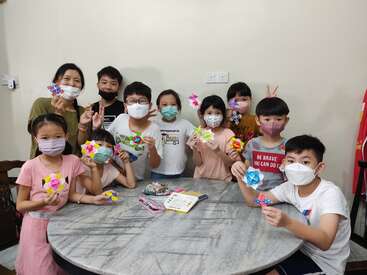 Un grupo de niños y un adulto, todos con mascarillas, muestran con orgullo coloridas manualidades de origami mientras se reúnen alrededor de una mesa redonda de mármol, sonrientes y alegres.