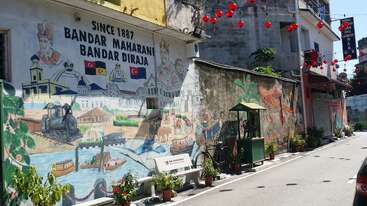 Un vibrante mural callejero en Bandar Maharani, Malasia, representa escenas históricas, la cultura local y lugares emblemáticos. Por encima cuelgan faroles rojos, y junto a ellos hay macetas con plantas y objetos vintage.