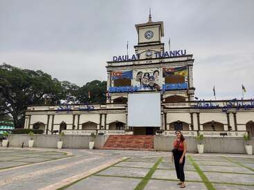 Una mujer está de pie frente a un impresionante edificio histórico con una torre del reloj, grandes carteles, murales, banderas y frondosos árboles bajo un cielo nublado.