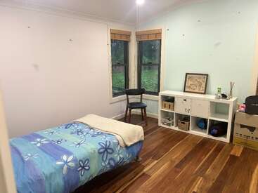 Un dormitorio ordenado con suelo de madera, cama individual con manta de flores azules, dos ventanas, estantería de cubos blancos, obras de arte, silla y algunas cajas en un rincón.