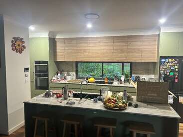 Esta imagen muestra una cocina moderna con armarios verdes y de madera, una isla de mármol, taburetes, frutero, una gran ventana y diversos electrodomésticos y adornos de cocina.