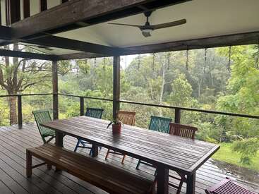Un balcón de madera con una larga mesa, bancos, sillas de colores, ventilador de techo y exuberantes vistas al bosque verde, que crean un espacio tranquilo y relajante para comer al aire libre.