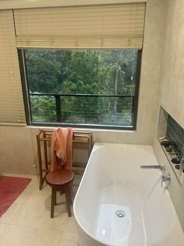 Un cuarto de baño moderno con bañera blanca, taburete de madera, toallero y toalla rosa, con un gran ventanal con vistas a frondosos árboles verdes, que crea un ambiente tranquilo.