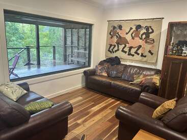 Esta acogedora sala de estar cuenta con sofás de cuero marrón, almohadas de colores, suelo de madera, arte tribal en la pared, una gran ventana y una vista panorámica de la vegetación exterior.