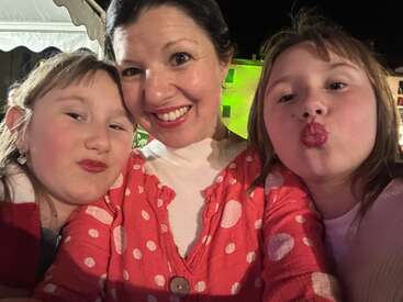 A woman and two children take a cheerful selfie at night. All are smiling brightly, with the children making playful faces. Everyone wears red lipstick.
