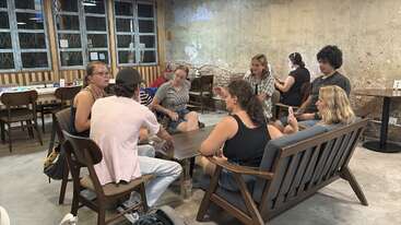 Eine Gruppe von Menschen sitzt um einen Holztisch in einem gemütlichen, rustikalen Café. Sie sind in ein Gespräch vertieft und scheinen sich zu amüsieren.