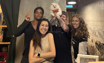 Vier Freunde posieren fröhlich im DeCafe Height & Seek. Einer von ihnen hält einen flauschigen weißen Hund hoch. Alle lächeln und verbreiten eine freundliche und fröhliche Stimmung.