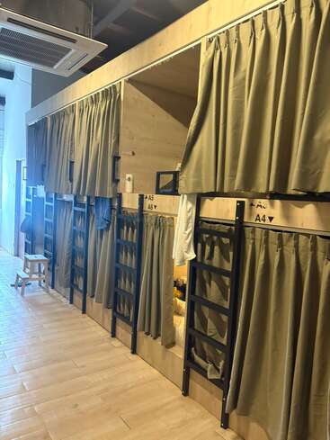 Das Bild zeigt eine Reihe gemütlicher Kapselbetten mit Sichtschutzvorhängen, Leitern und Holzboden, wahrscheinlich in einem modernen Hostel oder einer Gemeinschaftsunterkunft.