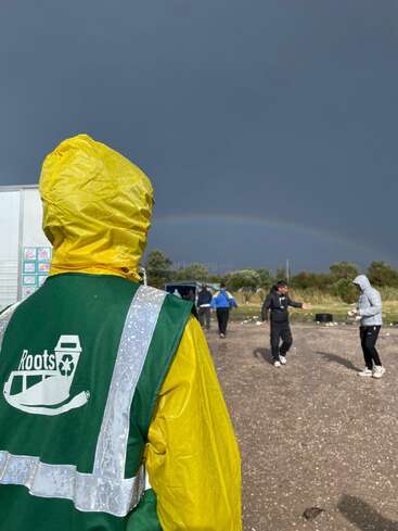 Eine Person in einem gelben Regenmantel und einer grünen Weste steht im Freien. Mehrere andere gehen in der Nähe. Ein schwacher Regenbogen erscheint unter einem dunklen, stürmischen Himmel.