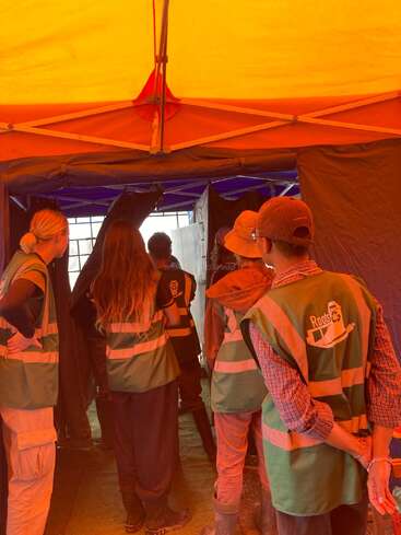 Eine Gruppe von Menschen mit grünen Warnwesten steht in einem Zelt, den Blick nach vorne gerichtet, und scheint Anweisungen zu hören oder etwas zu beobachten. Die Beleuchtung ist orange-gelb.