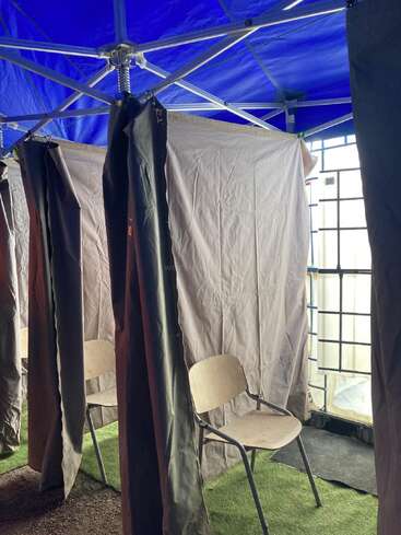 Dieses Bild zeigt eine behelfsmäßige Kabine mit einem Holzstuhl im Inneren, umgeben von Stoffvorhängen unter einem blauen Baldachin, die wahrscheinlich als private Kabine oder Station diente.