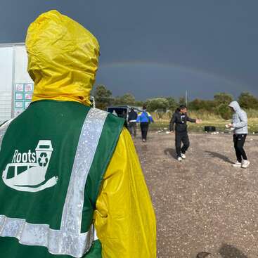 A person in a yellow raincoat and green vest stands outdoors. Several others walk nearby. A faint rainbow appears under a dark, stormy sky.