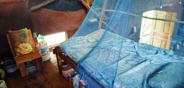 Cette chambre confortable dispose d'un lit avec des draps à motifs bleus et une moustiquaire, d'une chaise en bois avec des oreillers, d'une petite fenêtre et d'une porte partiellement ouverte.