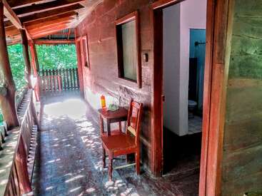 Un porche rústico de madera con una mesa y una silla, la luz del sol filtrándose entre los árboles, una puerta abierta que da a una pequeña habitación y otra puerta que conduce al cuarto de baño.