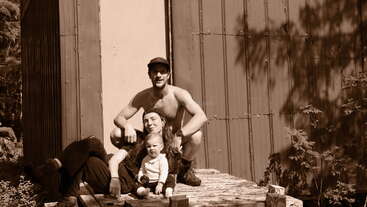 A sepia-toned photo shows a smiling shirtless man, a woman, and a small child sitting together outside near a rustic metal building, enjoying nature.