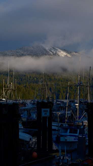 A peaceful harbor filled with boats sits in the foreground. Misty clouds drift above pine forests, with snow-dusted mountains glowing under a dramatic, brooding sky.