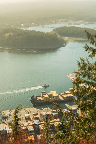 A container ship docked at a port, surrounded by colorful cargo containers. A small tugboat sails nearby, while forested islands and calm water create a serene atmosphere.