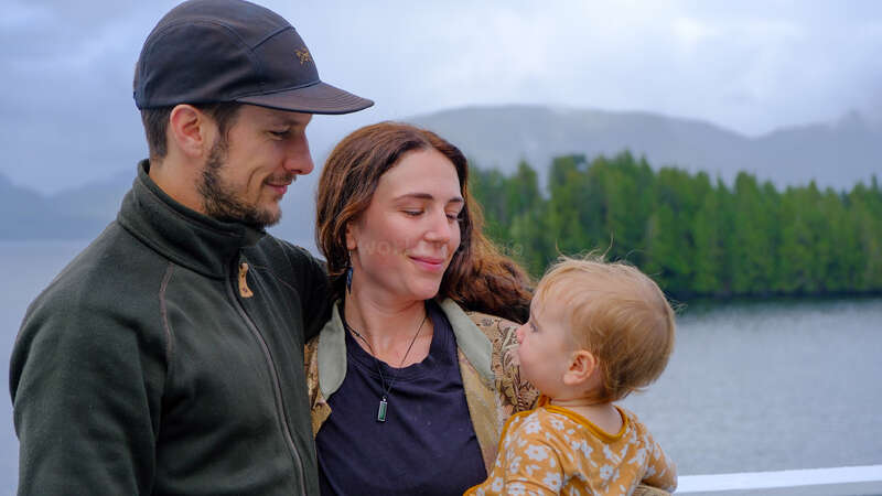 A happy family stands together outdoors, embracing each other. Behind them, lush green trees and a calm lake create a peaceful, scenic background with mountains and clouds.