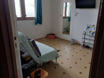 Una habitación modesta con un futón a rayas, suelo de baldosas, un espejo grande, televisión, una pequeña estantería, una papelera, ventanas con cortinas azules y algunos objetos personales esparcidos.