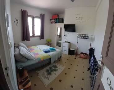 Um quarto aconchegante com cama, colcha colorida, travesseiros e toalhas. Tem uma TV na parede, cômoda, espelho, cabideiro e janela com cortinas. É bem iluminado e arrumado.
