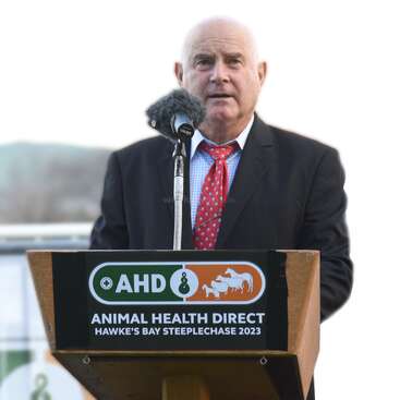 A bald man in a suit and red tie speaks at a podium labeled "AHD Animal Health Direct Hawke's Bay Steeplechase 2023" with a microphone.